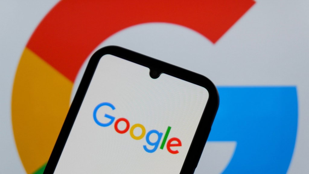 Celula com letreiro do Google na tela; ao fundo, tela maior exibe logomarca da empresa