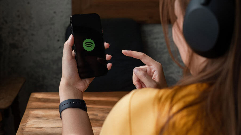 Jovem de costas com fones mexendo no app Spotify em um celular.
