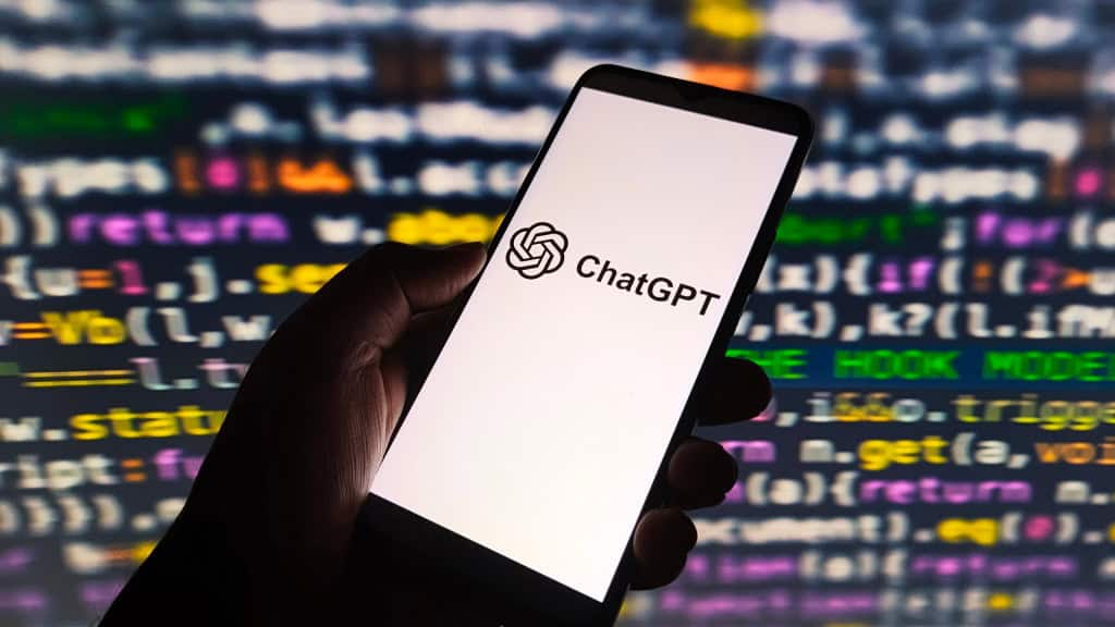 Ao fundo, tela com códigos de programação; à frente, mão de uma pessoa segurando um celular que exibe o logo do ChatGPT