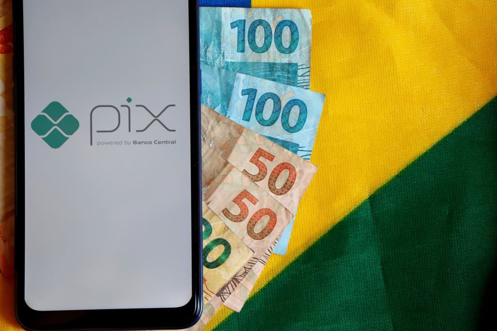 Celular exibe a tela inicial do Pix ao lado de cédulas de real sobre uma bandeira do Brasil.