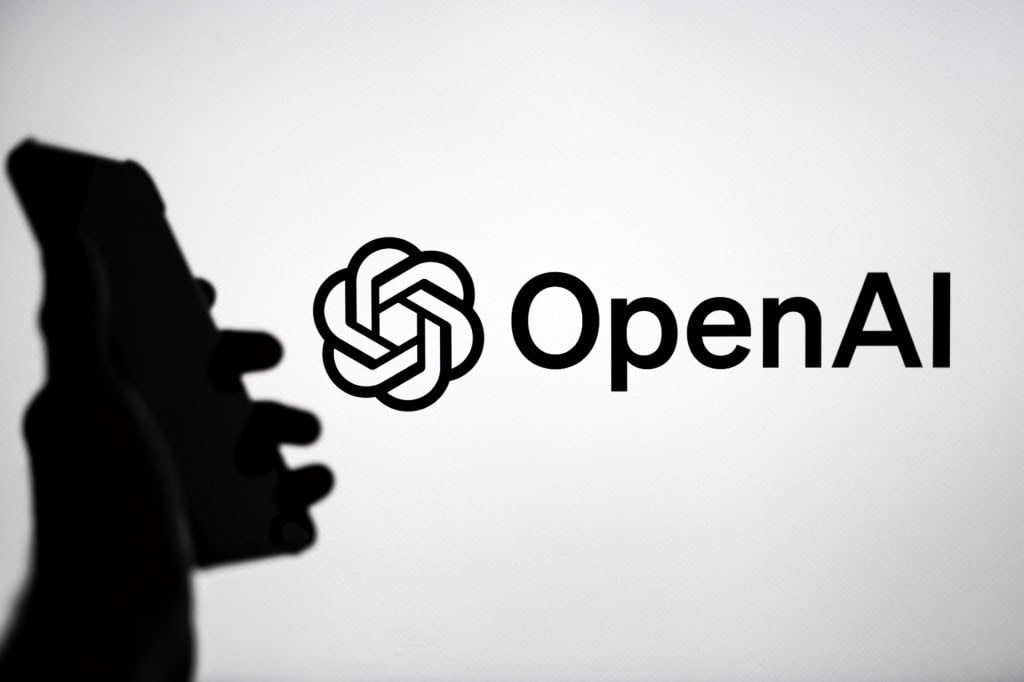 Ao fundo, logo da OpenAI; à frente e a esquerda, sombra de uma mão segurando um smartphone