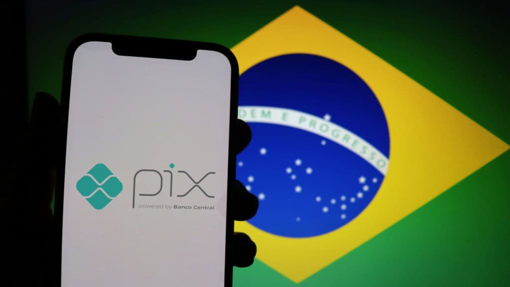 Pessoa segura celular exibindo logomarca do Pix na tela; ao fundo, monitor exibe bandeira do Brasil
