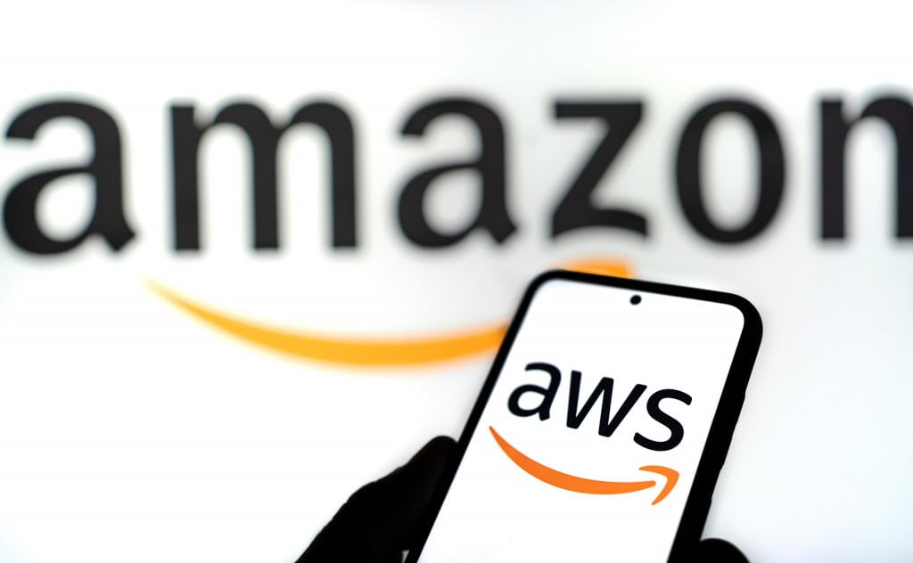 Ao fundo e parcialmente desfocado, o logo da Amazon; à frente, logo da AWS em um smartphone